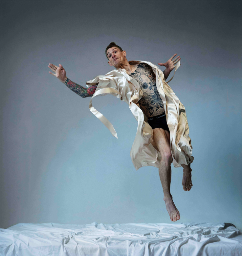 A tattooed, barefoot person is captured mid-jump above a white bed, their pale robe billowing as they float against a soft grey studio backdrop.