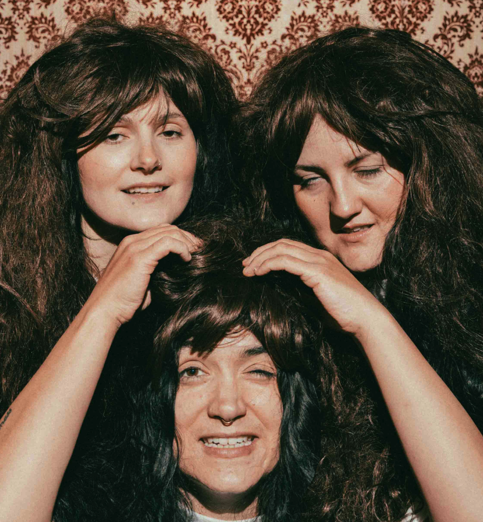 Three performers with long dark wigs pose closely together against patterned wallpaper, styled like the band The Shaggs.