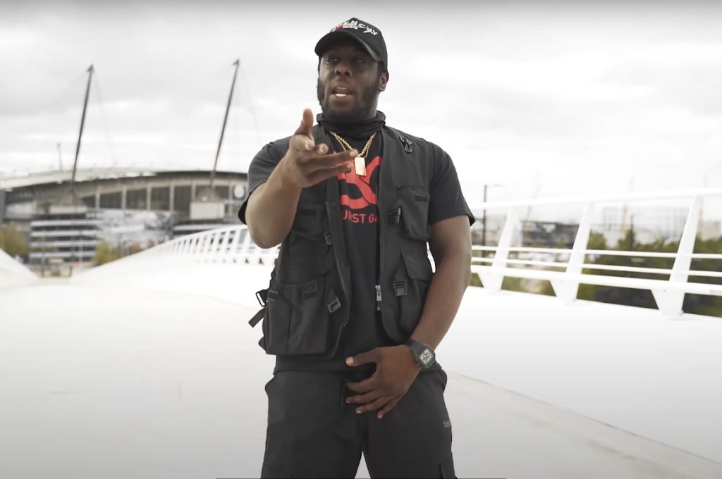 A man in black streetwear stands on a bridge, gesturing as he performs. A large stadium rises in the background under an overcast sky.