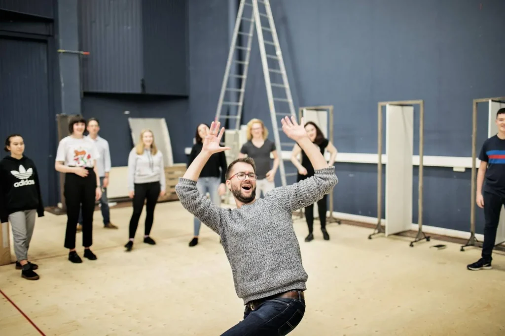 A man gestures energetically with raised arms in a rehearsal space, leading a group of people standing behind him. The room is spacious, with ladders and stage equipment in the background.