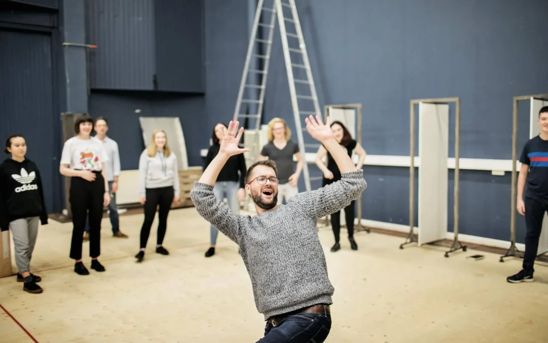 A man gestures energetically with raised arms in a rehearsal space, leading a group of people standing behind him. The room is spacious, with ladders and stage equipment in the background.