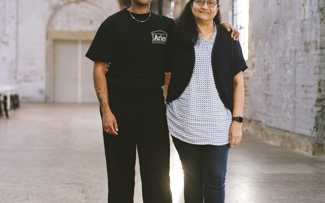 Two people stand side by side in a large, softly lit industrial hall with high ceilings and textured walls. One wears a red cap, black T-shirt, and black trousers, with a relaxed, confident stance and an arm around the other person. The second person wears glasses, a patterned top, dark cardigan, and jeans, smiling warmly. Light streams in from tall windows behind them, creating a bright strip on the floor between them and highlighting a calm, intimate moment of connection.