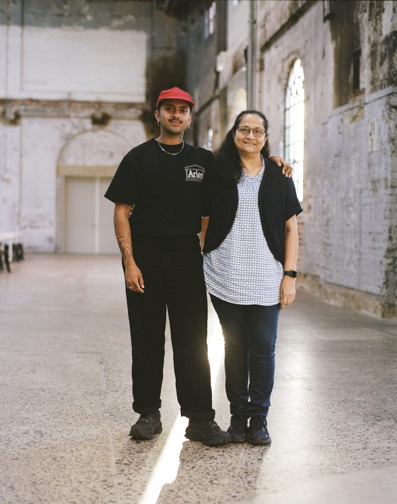 Two people stand side by side in a large, softly lit industrial hall with high ceilings and textured walls. One wears a red cap, black T-shirt, and black trousers, with a relaxed, confident stance and an arm around the other person. The second person wears glasses, a patterned top, dark cardigan, and jeans, smiling warmly. Light streams in from tall windows behind them, creating a bright strip on the floor between them and highlighting a calm, intimate moment of connection.