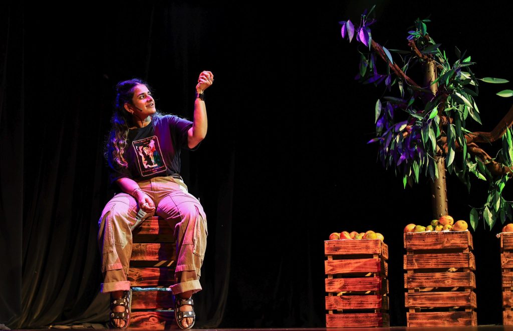 A performer sits on stacked wooden crates on a dimly lit stage, gesturing upward. Beside them are crates of fruit and a small tree, illuminated by warm, colourful stage lighting.