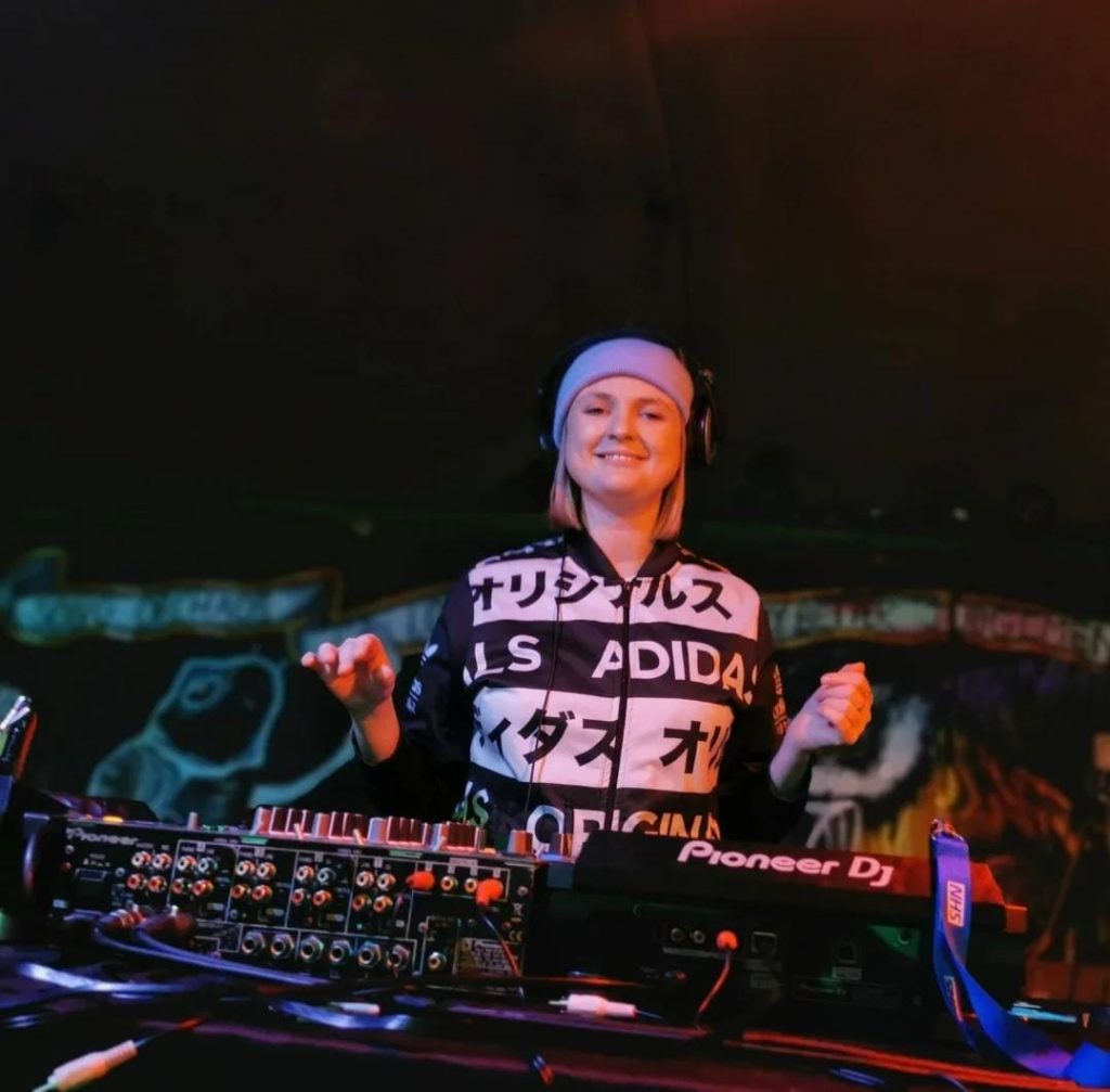 The image shows a person DJing at what looks like a club or live music venue. They’re standing behind a DJ setup (including a Pioneer DJ controller) and wearing headphones and a black-and-white patterned top with bold text. They’re smiling and appear relaxed, possibly mid-performance or enjoying the music. The lighting is low with warm reddish/orange tones, and there’s graffiti-style artwork visible in the background, which gives the space an underground or creative vibe. Their hands are slightly raised, as if they’re feeling the beat or about to adjust controls. Overall, the scene captures a lively, intimate DJ performance moment with a confident, upbeat mood.