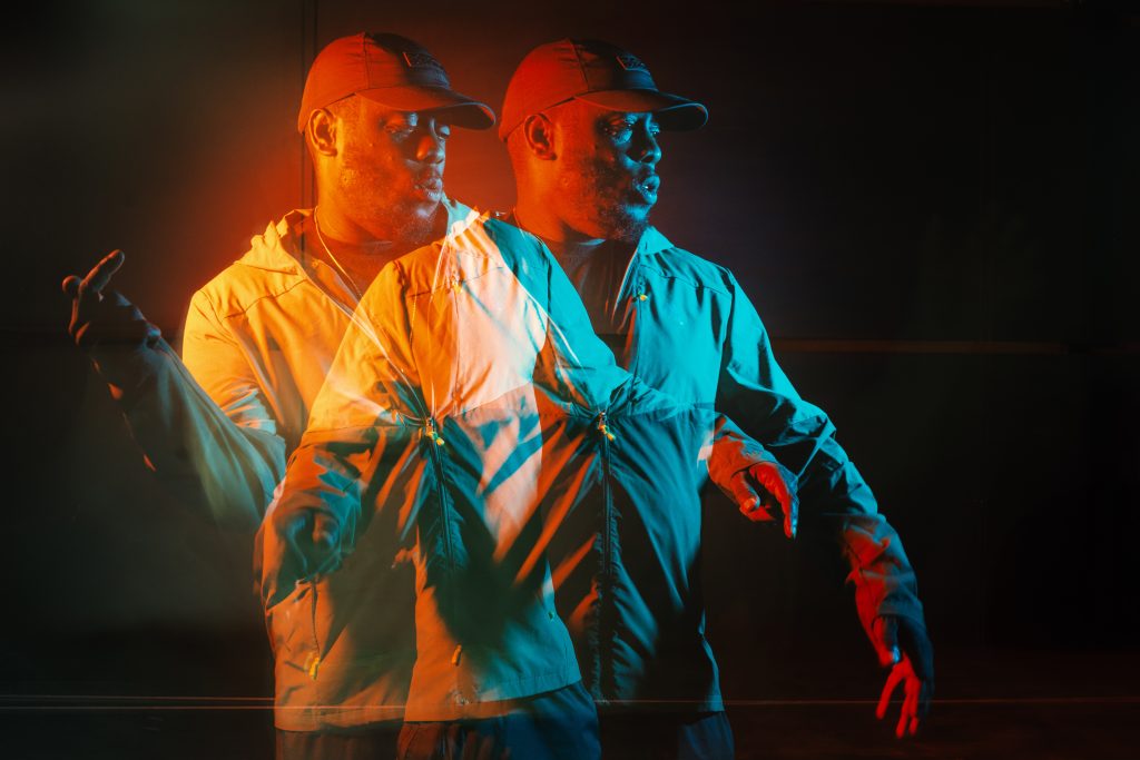 A performer (Hypes) in a baseball cap and light tracksuit is captured in motion using a double-exposure effect. Dramatic red-orange and blue lighting creates two overlapping figures against a dark background, suggesting movement and performance.