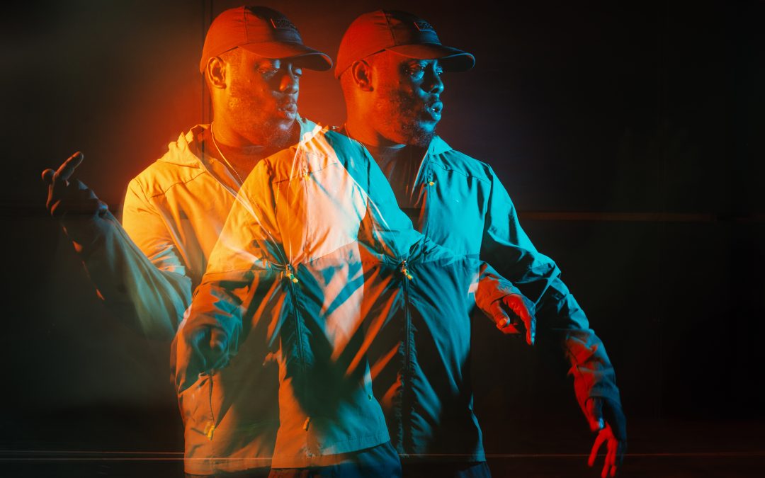 A performer (Hypes) in a baseball cap and light tracksuit is captured in motion using a double-exposure effect. Dramatic red-orange and blue lighting creates two overlapping figures against a dark background, suggesting movement and performance.