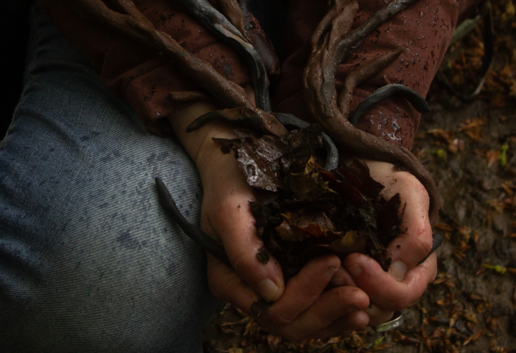 Close-up of muddy hands holding wet leaves and forest soil, intertwined with dark, root-like branches, against an earthy woodland background.